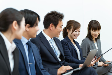 会社でパソコン・スマホ・タブレットを使うスーツ姿の会社員（アジア人・男女・5人）
