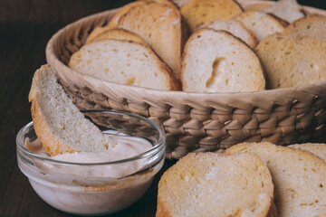 Pieces of roasted white bread in wicker basket and glass bowl of sauce on a dark background. Closeup of delicious croutons.
