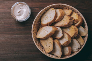 Pieces of roasted white bread in wicker basket and glass bowl of sauce on a dark background. Closeup of delicious croutons. Top view.