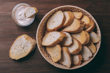 Pieces of roasted white bread in wicker basket and glass bowl of sauce on a dark background. Closeup of delicious croutons. Top view.