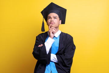 Thoughtful face of young male wearing graduation cap and ceremony robe over yellow background with...