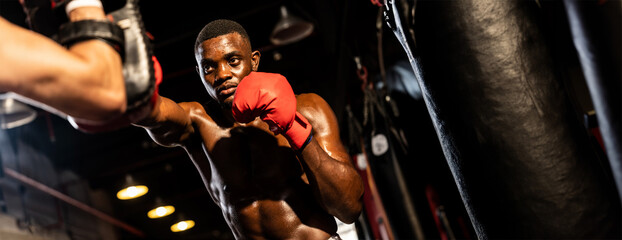 African boxer punching at trainer or coach wearing punching mitts as boxing bag training equipment in the gym. Strength and stamina training for professional boxing match. Spur