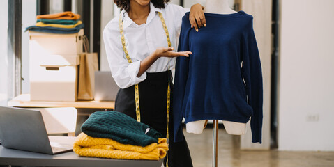 Female fashion, designer, Looking at Drawings and Sketches that are Pinned to the Wall Behind Her Desk. Studio is Sunny. Personal Computer, Colorful Fabrics, Sewing Items are Visible.