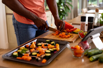 Midsection of african american man chopping vegetables using tablet in kitchen