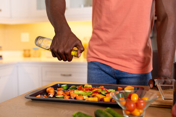 Midsection of african american man pouring oil on vegetables in kitchen