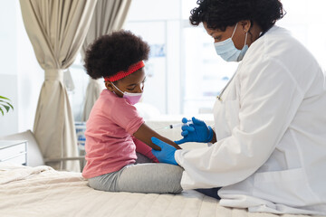African american female doctor wearing face mask vaccinating girl patient at hospital