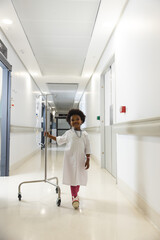 Portrait of happy african american girl patient holding drip and walking in corridor at hospital