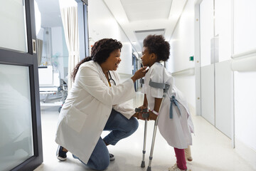 African american female doctor and girl patient walking with crutches in corridor at hospital