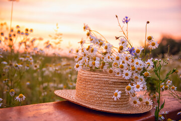 A straw hat and a bouquet of wild flowers on a wooden bench in a field at sunset on a summer evening. - 626160314