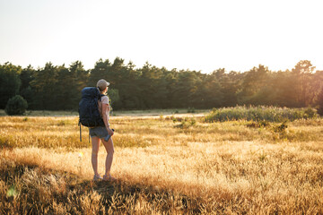 Naklejka na ściany i meble Female hiker with backpack and thermos is resting in nature and enjoying summer sunset. Woman relax while hiking on a long trekking trail