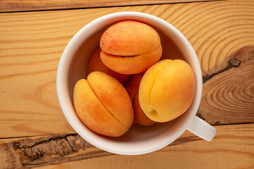 Several ripe juicy apricots in a white ceramic cup on a wooden table, macro, top view.
