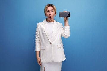 young surprised well-groomed slender blond woman wearing a white jacket and dress holds a smartphone horizontally to insert an advertising offer