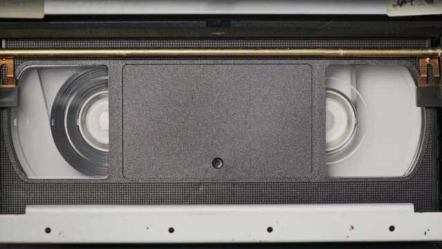 VHS cassette is played inside a VCR tape recorder, top view. Videotape reels rotate. Videocassette with empty blank tag is starting playback. Old video recorder inside close-up. Playing vintage movie
