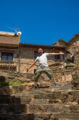Bald man with half a beard smiling with open mouth, expressive eyes and arms outstretched to give a cheerful and funny surprise standing on a stone staircase in a rural village on a sunny day
