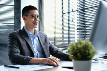 Elegant Asian businessman wearing glasses looking at computer monitor, doing online data market analysis in modern workplace