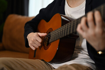 Cropped shot of man playing acoustic guitar on couch, spending leisure time in cozy living room