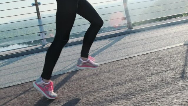 Crop woman legs in dark leggings and sneakers with pink shoelace sprinting across bridge. Skinny female runs on asphalt city bridge