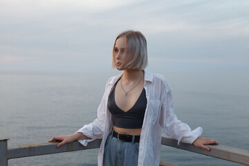 Portrait of  blonde woman in white shirt and tank top standing on the background of sea, cloudy sky