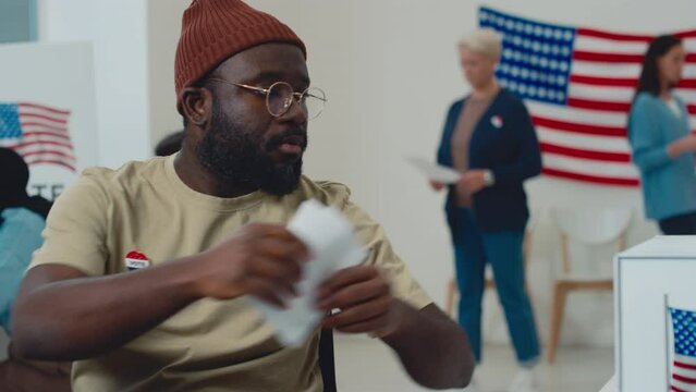 Medium shot of middle-aged African American man in wheelchair, wearing casual beanie and glasses, riding up to ballot box with USA flag emblem at electoral station, casting his bulletin and leaving