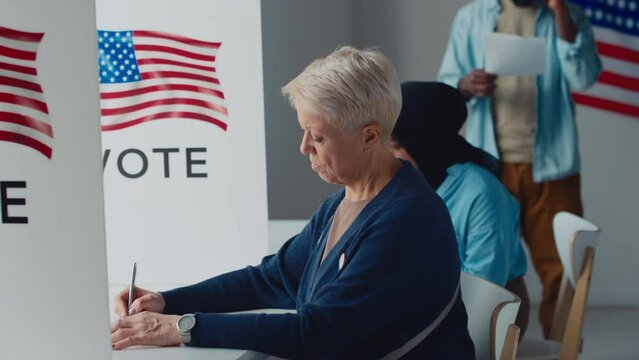 Medium side shot of middle-aged Caucasian lady and Muslim woman sitting in booths at polling station, writing on ballot and leaving, diverse American citizens queuing, USA flag emblem in background