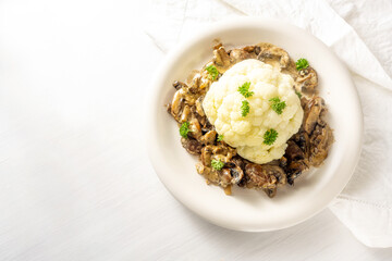 Cauliflower with mushroom ragout and parsley garnish on a plate and on a white table, healthy vegetable meal, high angle view from above, copy space, selected focus