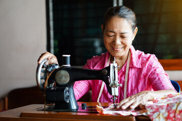 Happy senior Asian woman tailor is sewing cloth by vintage sewing machine at home. Concept, Senior  lifestyle, spend  free time to repair or design clothes. Hobby, not lonely.                         