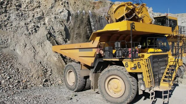 Large bucket excavator loads ore into a large dump truck in open mine gold digging pit, slow motion aerial shot