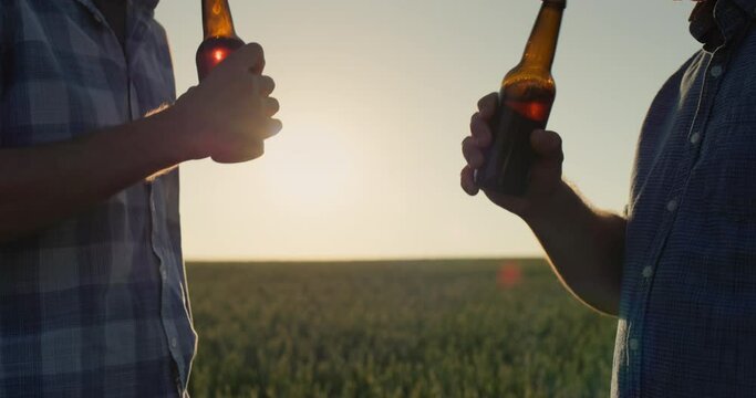 Two farmers clink bottles of beer against the background of a field of wheat, only hands are visible in the frame