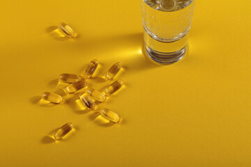 Diet, nutrition, healthy eating concept. Oil filled capsules of food supplements and glass of water for pouring yellow softgels capsules on yellow background.