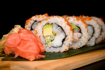 Closeup view of California rolls covered orange tobiko with red and white ginger nearby served on banana leaves on wooden board