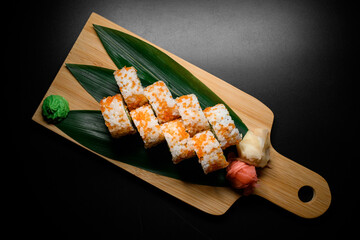 Appetizing California rolls covered with orange tobiko caviar with red, white ginger and wasabi nearby served on banana leaves on wooden citting board