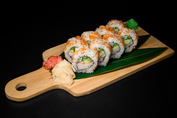 Side view of California rolls covered with orange caviar with red, white ginger and wasabi nearby served on banana leaves on wooden board. Black background.