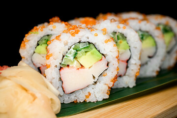 California rolls covered with orange tobiko caviar served on banana leaves on wooden board on black background.