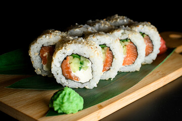 Side view of sushi roll with rice, cream cheese, cucumber, salmon, sesame served on green banana leaves and wooden board