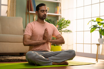 Indian young man doing namaste posture or yoga with closed eyes while sitting at home - concept of healthy lifestyle, fitness and self caring - obrazy, fototapety, plakaty