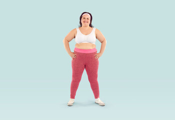 Happy funny young fat woman in sportswear looking at camera standing confidently with hands on the sides isolated on a studio blue background ready for sport fit exercises. Workout, gym concept.