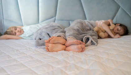 Two barefoot children lie side by side under a blanket on a large bed. Selective focus on the bare...