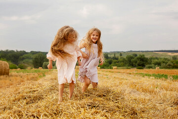 Naklejka na ściany i meble two long-haired blonde girls in linen dresses jump on a haystack in a mown wheat field