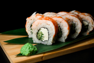 Pieces of sushi roll with shrimp served on wooden board with plant leaves and wasabi on black background