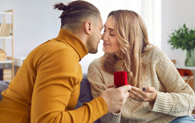 Young man with a red gift box proposing to his happy girlfriend sitting on the sofa. Lovely handsome guy giving surprise gift with a engagement ring or jewelry to his happy girlfriend.