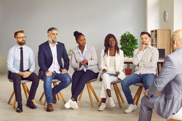Diverse team of people talking to professional business coach or corporate manager. Group of young and mature multiethnic men and women sitting in row in office and having discussion with senior man