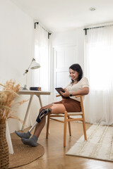 Woman with prosthetic leg  sitting in chair in living room at home. Relaxed, happy woman at home living room sitting on the chair, using tablet