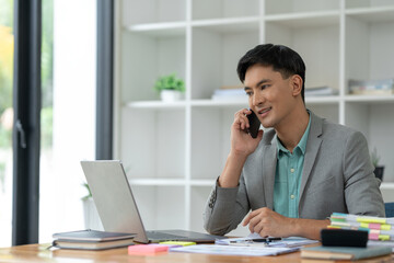 Happy young asian businessman looking at data on laptop, chatting on phone using smartphone for communication. Check through the application Go online while at work in the office.