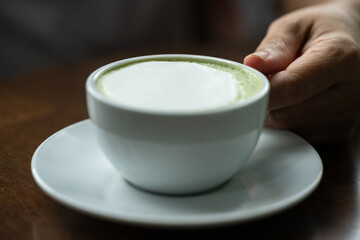 Closeup hand holding white cup of hot green tea latte on wood background in restaurant.Best of menu in the coffee shop.Soft focus.