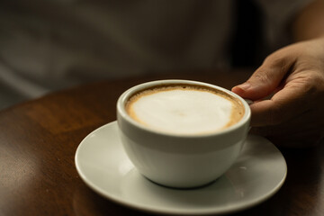 Closeup hand holding white cup of hot coffee latte on wood background in restaurant.Best of menu in...