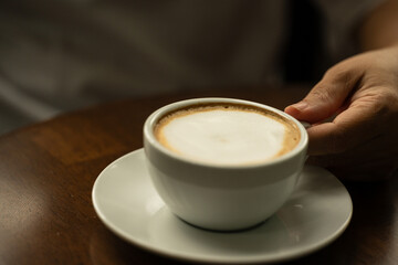 Closeup hand holding white cup of hot coffee latte on wood background in restaurant.Best of menu in the coffee shop.Soft focus.