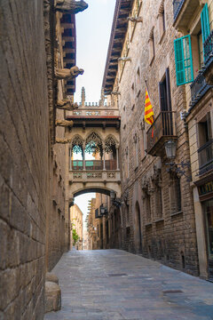 Daytime photo of the Pont del Bisbe or Bishop Street Bridge, in the Gothic Quarter of Barcelona, Spain. The Catalan flag is hanging from a balcony next to turquoise window shutters.