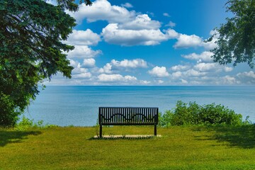 Under a partly cloudy blue sky on a Summer day, an empty bench faces the blue expanse of Lake Michigan from atop a wooded bluff near Milwaukee, WI. - Powered by Adobe