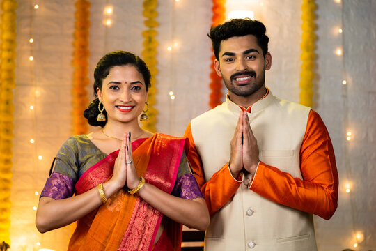 Happy young indian couple in traditional ethnic clothing saying namaste or welcome gesture by looking camera during festival celebration at home - concept of greeting, wishes and togetherness.