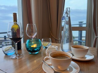 Coffee on the terrace of a restaurant with a beautiful view of the sea, with cups saucers with beverages, wine bottle, water drink and glasses on wood table looking out to ocean on light early evening